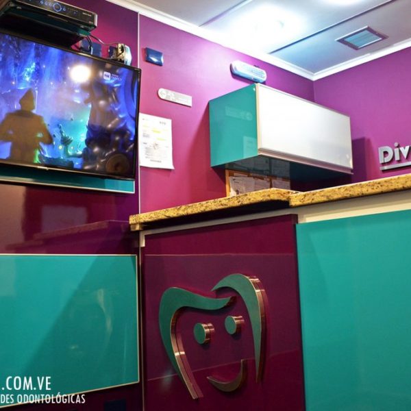 Mostrador de recepción de Divina Sonrisa en tonos turquesa y violeta con televisor y el logo de la clínica