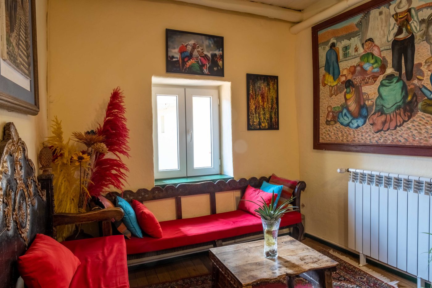 Sala de estar colonial del Hostal Corihuasi con sofá tallado, cojines de colores y pinturas costumbristas cusqueñas