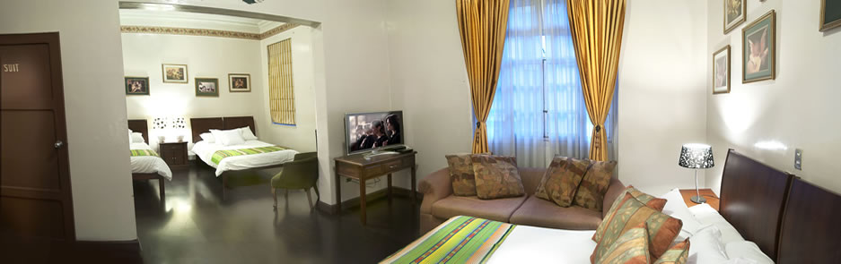 Suite del Hotel Cuenca con dos camas, sofá y televisión, decorada con muebles de madera colonial